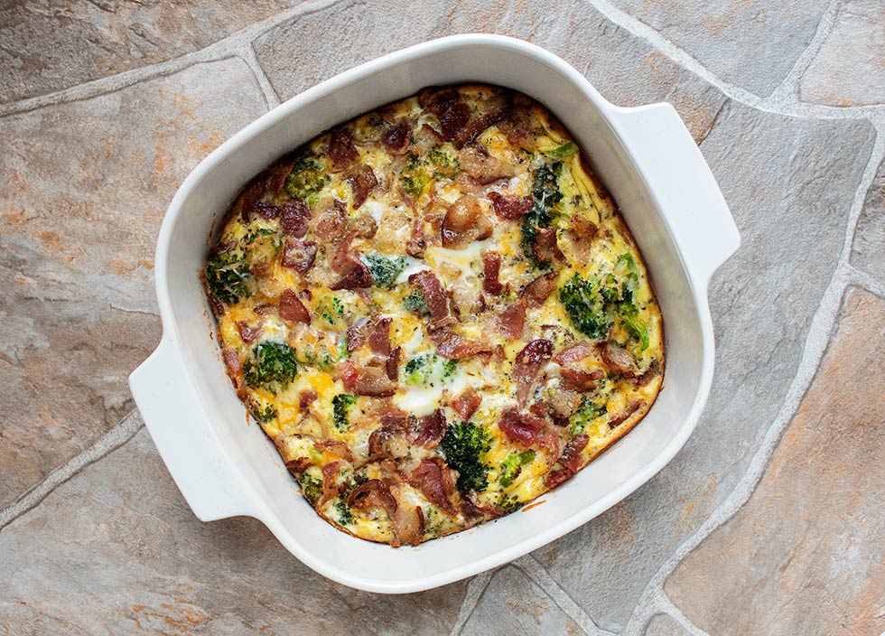 overhead view of breakfast casserole with eggs, bacon, broccoli, and melted cheese in a white casserole dish, placed on countertop, after it is removed from oven
