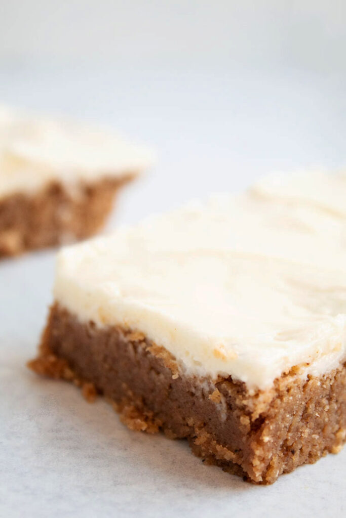 sideview of two brown pumpkin bars with white cream cheese frosting on top on a white countertop