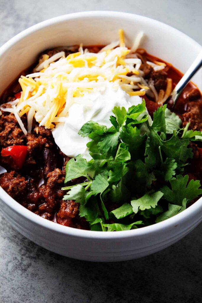 overhead view of white bowl with homemade beef chili inside and garnished with yellow and white cheese, green cilarntro, white sour cream on top and silver spoon