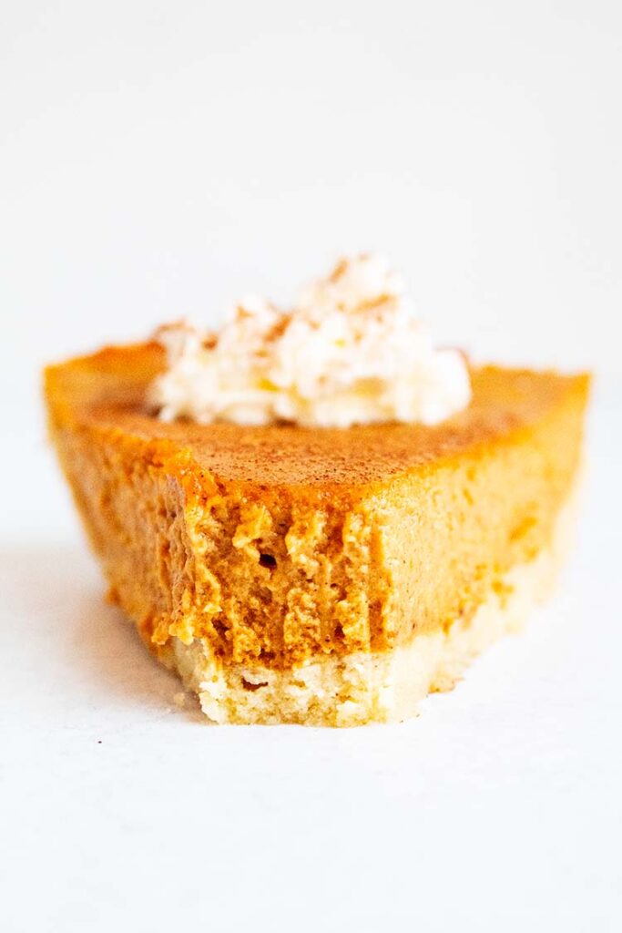 sideview of slice of pumpkin pie with whipped cream on top laying on a white countertop