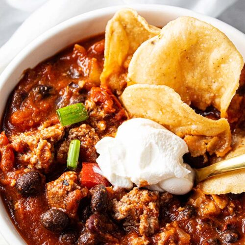 overhead view of turkey chili with dab of white sour cream , green onion garnish and a few tan chips in the upper right corner, all in a white serving bowl