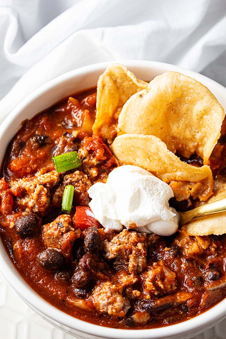 overhead view of turkey chili with dab of white sour cream , green onion garnish and a few tan chips in the upper right corner, all in a white serving bowl