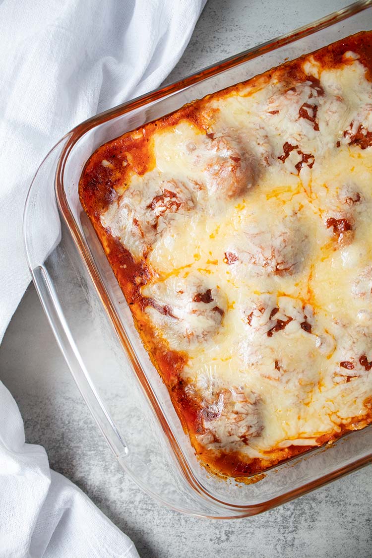 overhead view of meatball casserole topped with cheese in a glass casserole dish on a white marble countertop and clean white dishtowel laying next to it