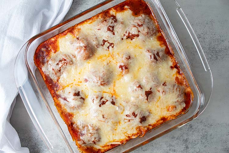 overhead view of meatball casserole topped with cheese in glass casserole dish on a white marble countertop and white dishtowel laying next to it