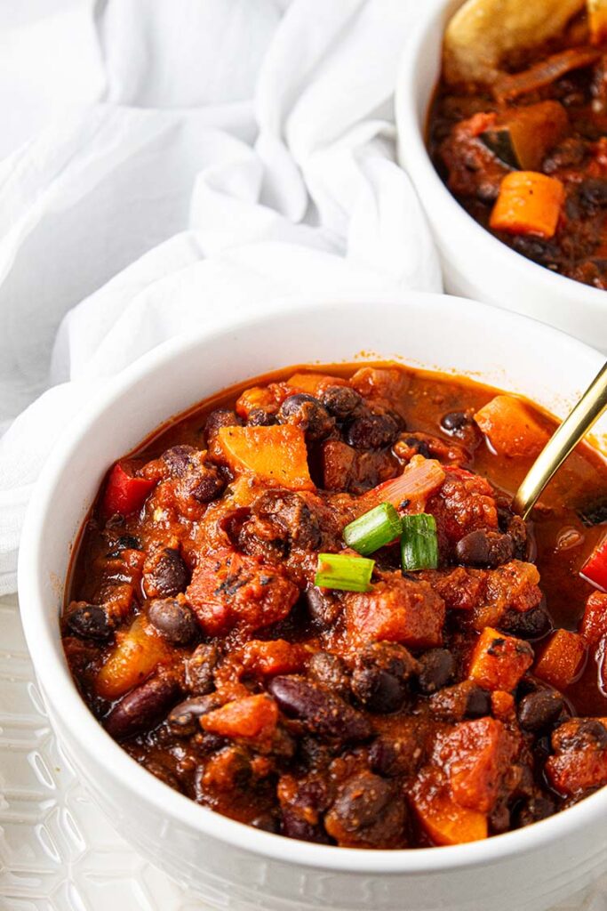 overhead view of vegetarian chili in white serving bowl garnished with green onions and with gold serving spoon next to second bowl of vegetarian chili