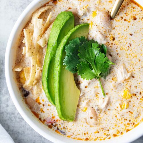 overhead view of white serving bowl with white chicken chili and avocado slices on top and gold spoon