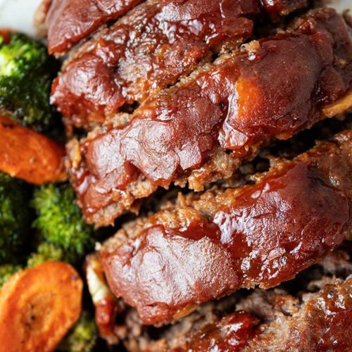 overhead view of sliced homemade glazed meatloaf topped with ketchup glaze and a side of roasted broccoli and carrots on a white serving dish