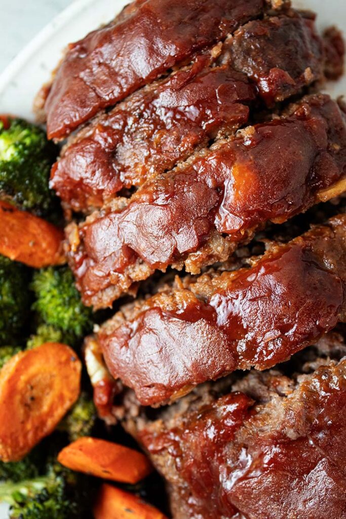 overhead view of sliced homemade glazed meatloaf topped with ketchup glaze and a side of roasted broccoli and carrots on a white serving dish