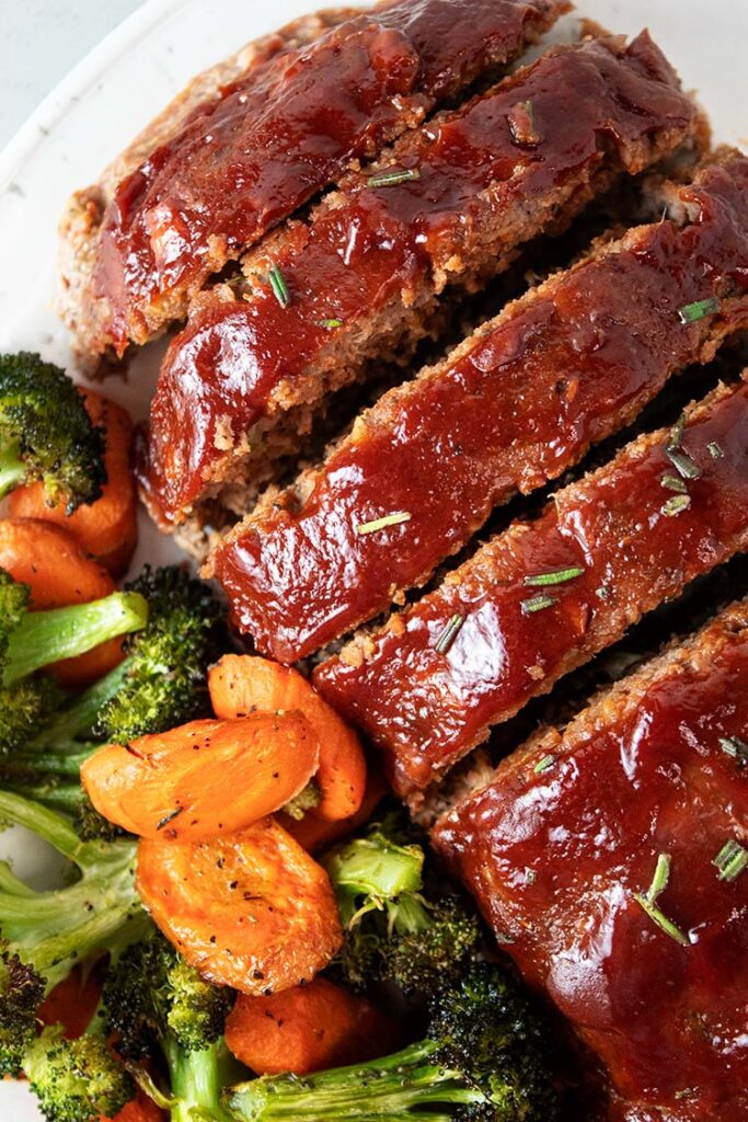 overhead view of turkey meatloaf on a white serving dish with ketchup glaze and healthy carrots and broccoli on the side