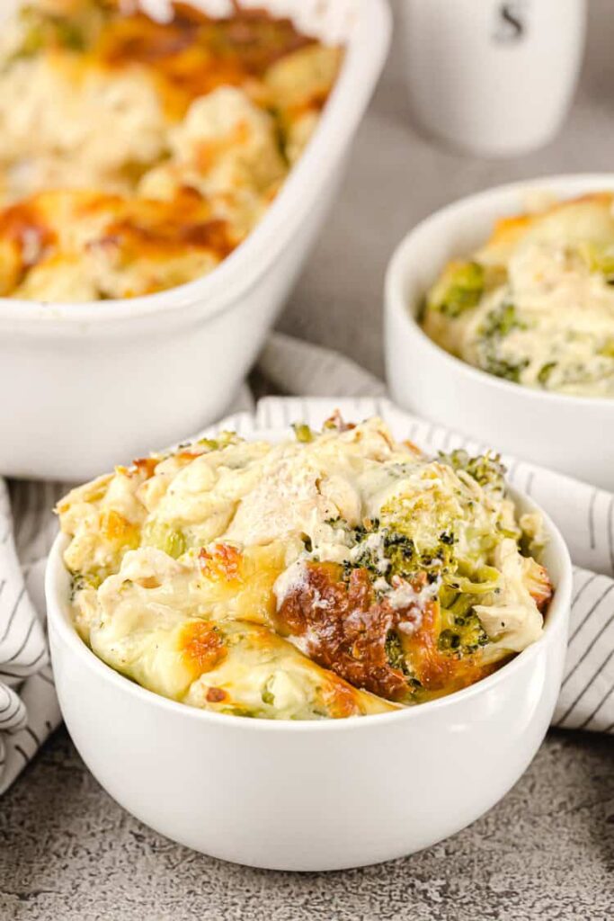 sideview of two white serving bowls of chicken and broccoli casserole inside and white casserole dish in background