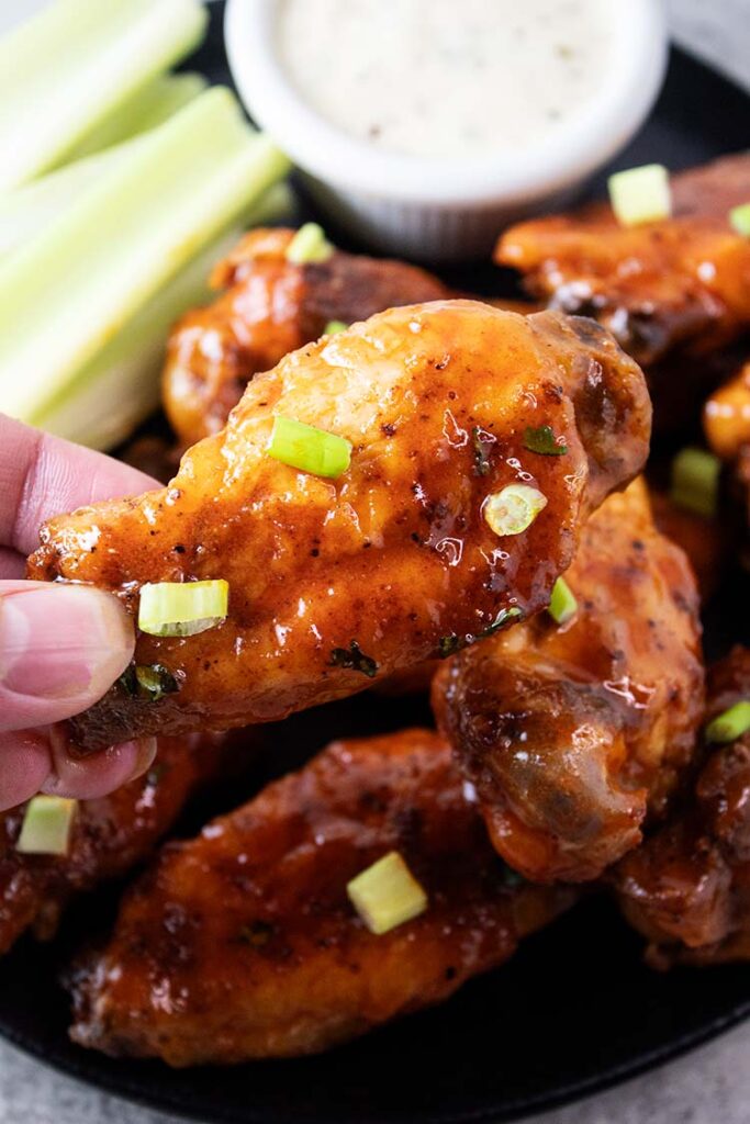 closeup view of fingers holding a hot wing with a black plate of air fryer buffalo hot wings with celery and side cup of blue cheese in the background