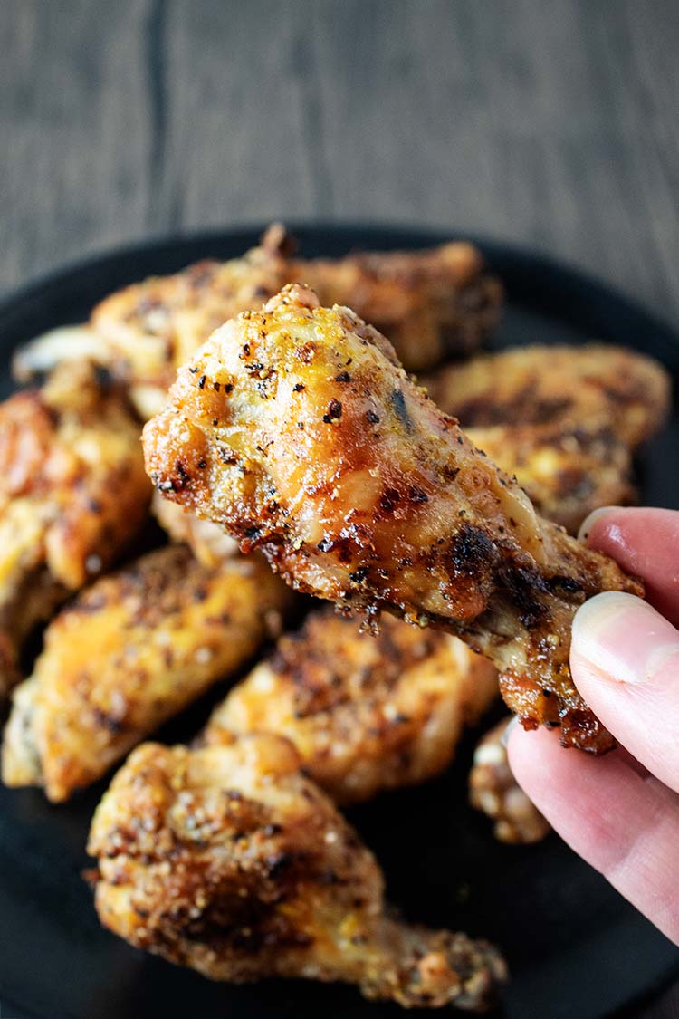 closeup view of person's hand holding a cooked wing in the foreground and a plate of air fryer lemon pepper wings in the background on a black serving plate