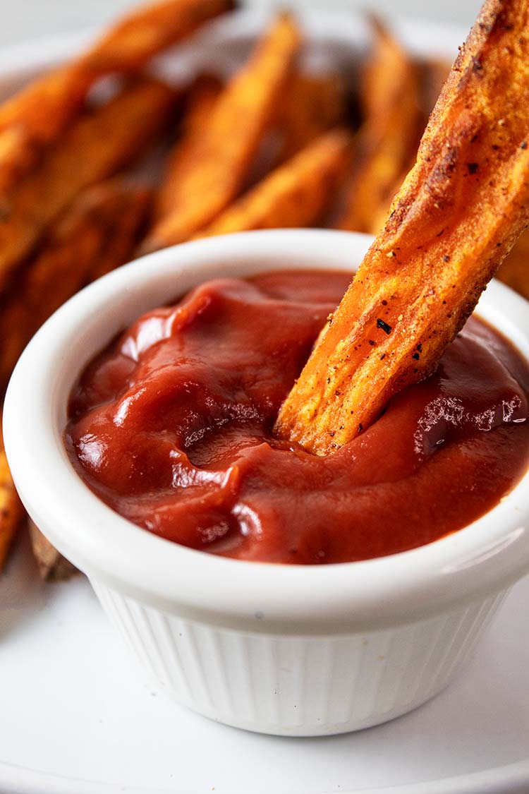 sideview of air fryer sweet potato fries on a plate in the background with one fry being dipped in a small white serving cup of ketchup