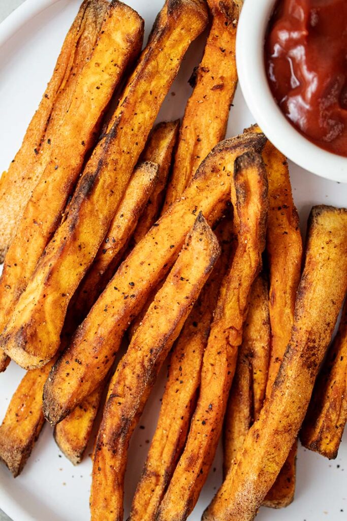 overhead view of air fryer sweet potato fries on a white serving plate with ketchup on the side in a white serving cup