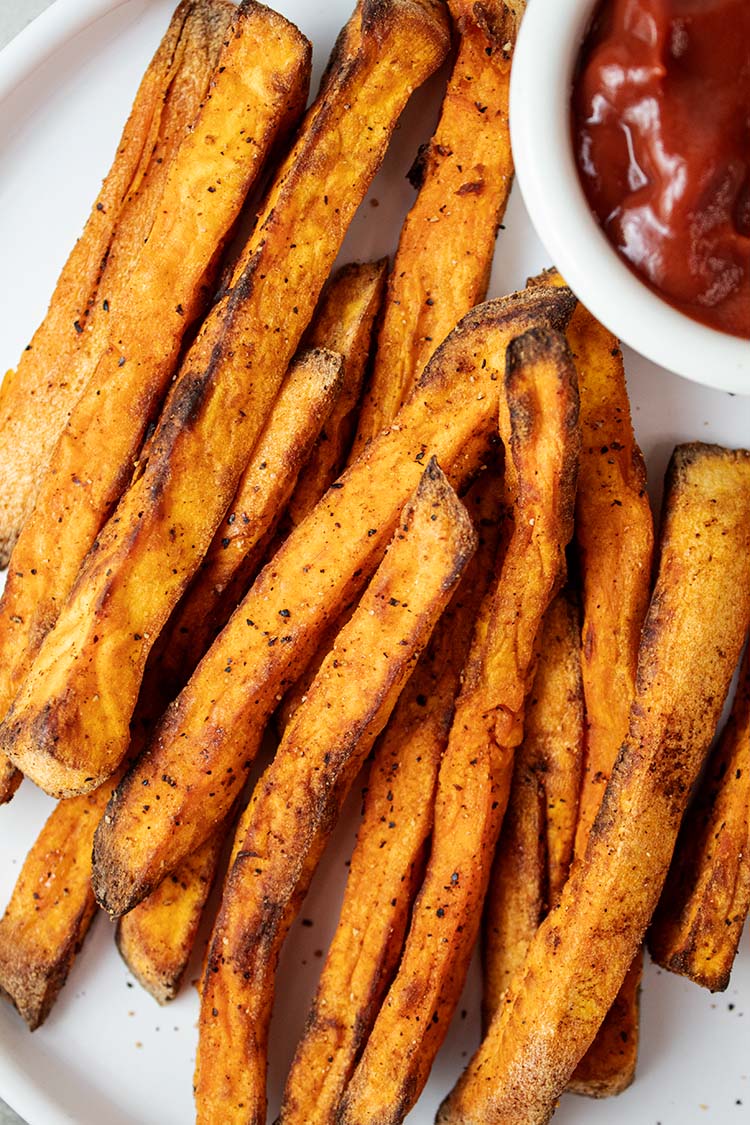 overhead view of air fryer sweet potato fries on a white serving plate with ketchup on the side in a white serving cup
