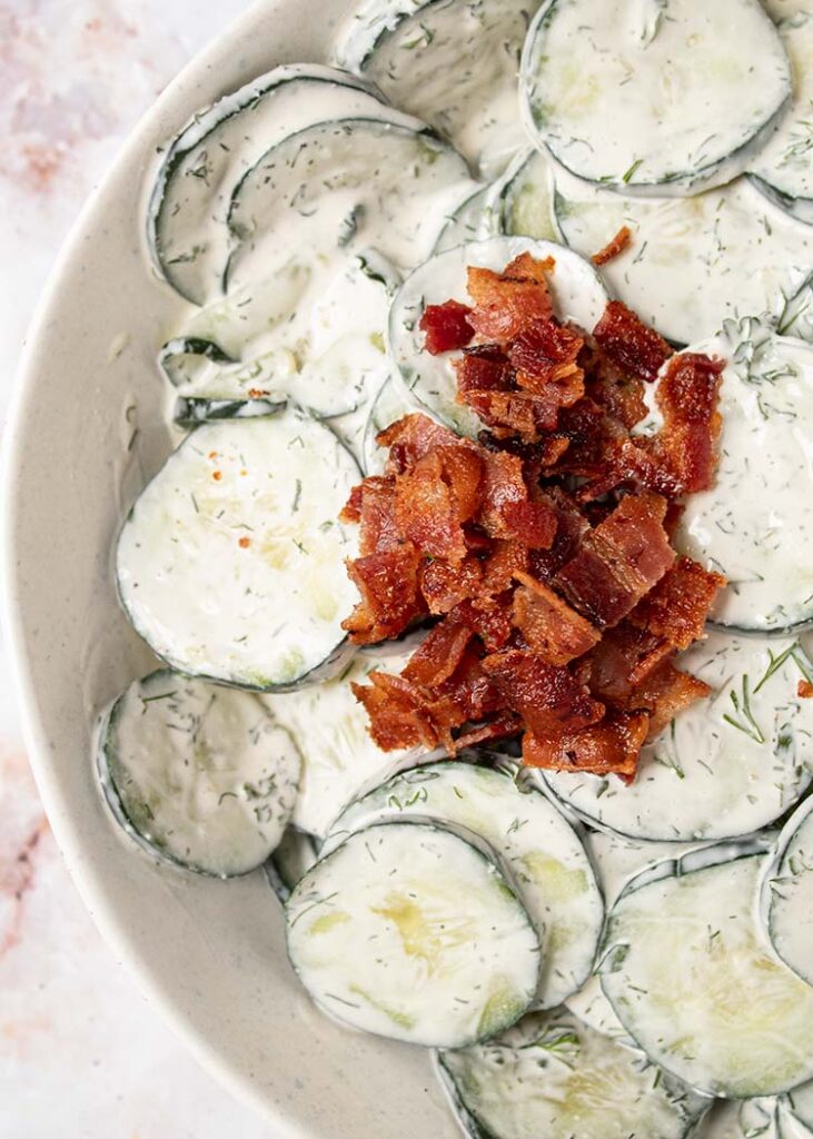 overhead view of cucumber ranch salad topped with bacon in a white serving bowl