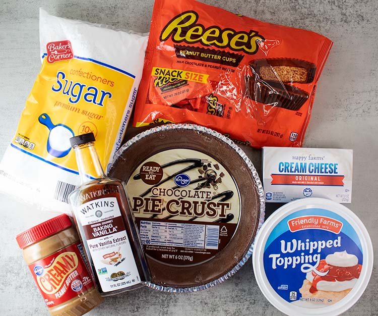 overhead view of ingredients for no bake peanut butter pie including chocolate grahram cracker crust, sugar, reeses cups, cream cheese, cool whip, vanilla and peanut butter