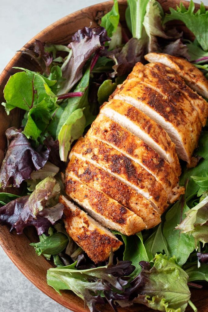 overhead view of a seasoned and evenly sliced air fryer chicken breast on a bed of salad greens in a wooden bowl, ready to be served