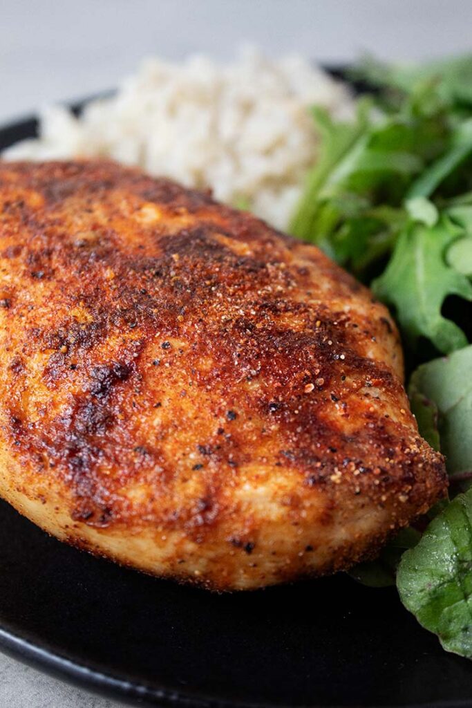 sideview of seasoned air fryer chicken breast on a black serving plate with sides of salad and white rice