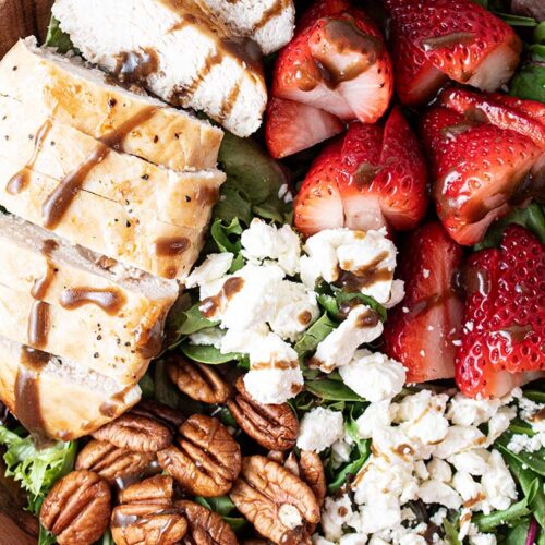 overhead view of ready to serve chicken and strawberry salad with sliced chicken, strawberries, pecans, feta cheese crumble all on a bed of lettuce in a wooden bowl
