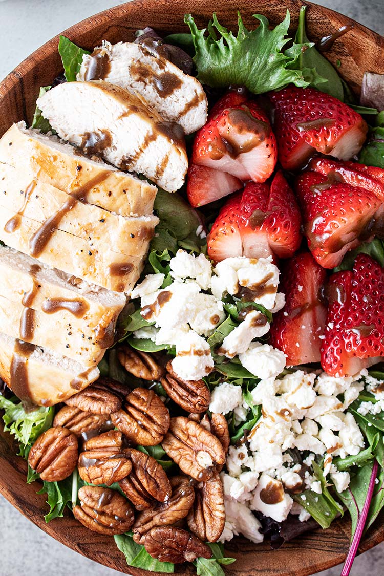 overhead view of ready to serve chicken and strawberry salad with sliced chicken, strawberries, pecans, feta cheese crumble all on a bed of lettuce in a wooden bowl