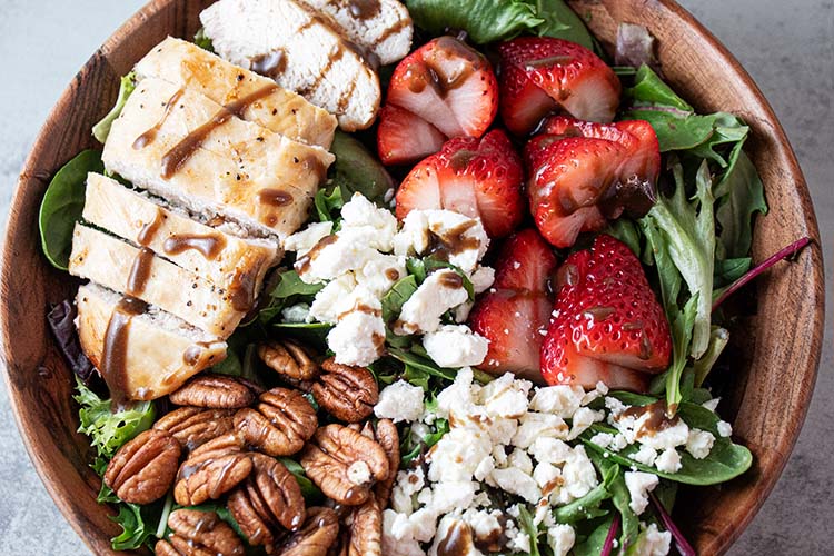 overhead view of ready to serve strawberry chicken salad with sliced chicken, strawberries, pecans, feta cheese crumble all on a bed of lettuce in a wooden bowl