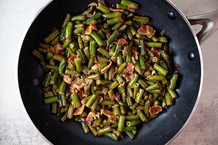 overhead view of bacon and green beans in black skillet during the cooking process
