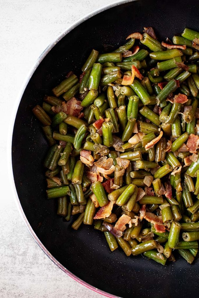 overhead view of cooked bacon green beans in a black skillet on a white background