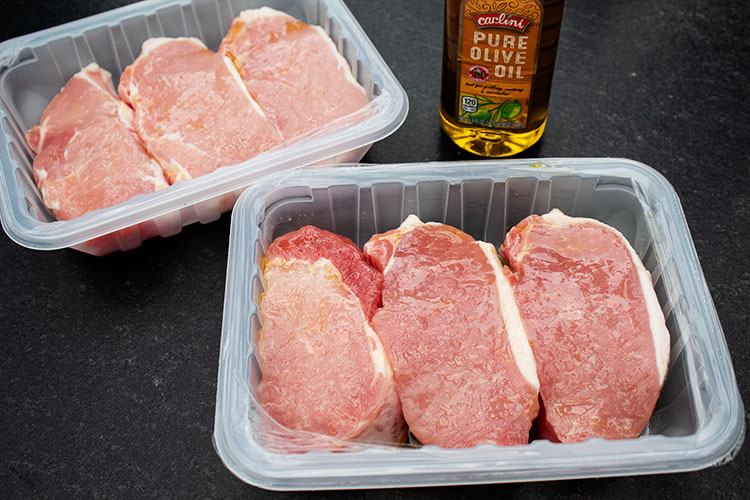 sideview of two packages of raw boneless pork chops with bottle of virgin olive oil next to them