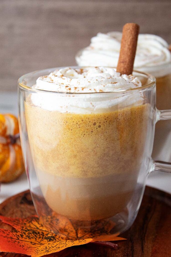 sideview of two ready to serve pumpkin spice latte coffee drinks in glass mugs topped with whipped cream and vanilla sticks and surrounded by fall decor