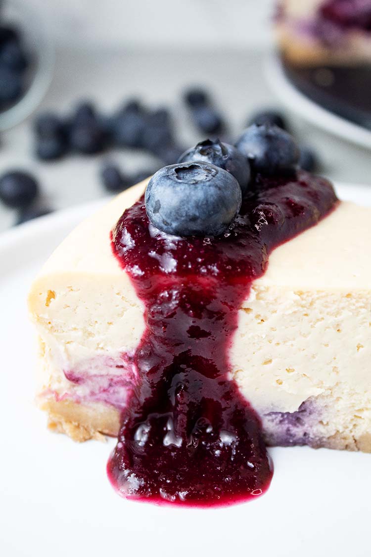 sideview of blueberry cheesecake ready to serve on a white dish topped with blueberry sauce and blueberries