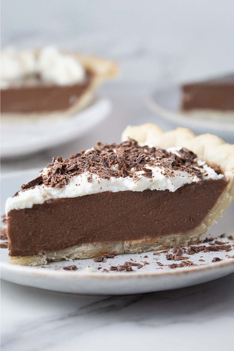 sideview of slice of chocolate pie topped with whipped cream and chocolate shavings on a white serving dish