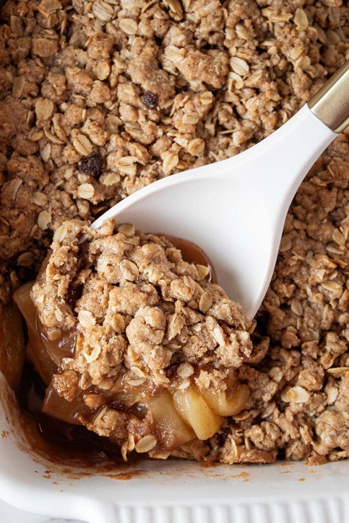 overhead view of apple crisp in a white casserole dish and a large white serving spoon removing one serving