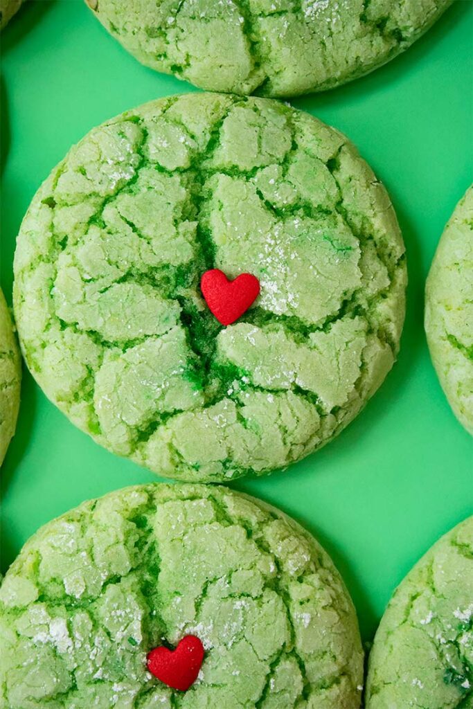 overhead view of cake mix grinch cookies with small decorative candy hearts in the center all lined up on a green surface and ready to serve