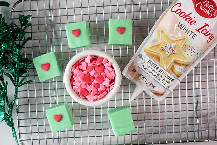 overhead view of grinch green fudge cut into squares with small red candy hearts added to the center of each and christmas decor supplies all laying on a wire rack