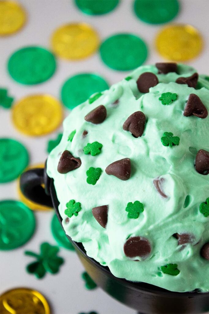 sideview of St. Patrick's Day cheesecake fluff dessert in a serving bowl topped with chocolate chips and mini shamock candies and decor all around the bowl