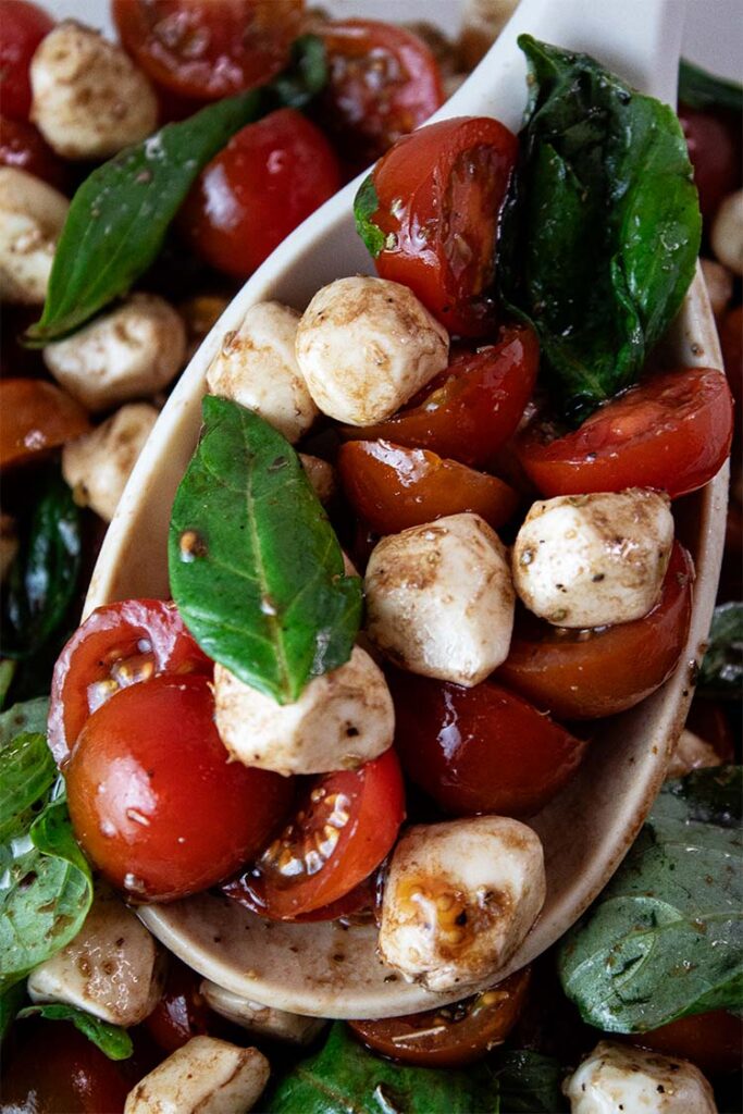 overhead view closeup of one serving of caprese salad on a serving spoon hovering over the entire glass serving bowl
