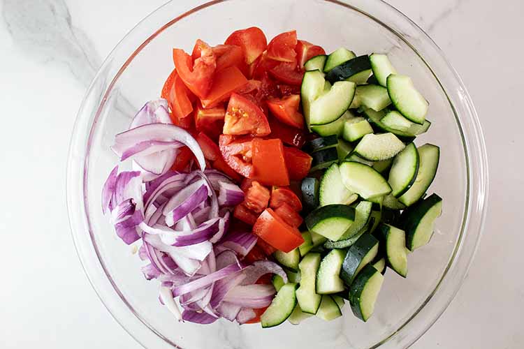 overhead view of glass mixing bowl with sliced red onions, chopped tomatoes and cucumber ready to be lightly tossed